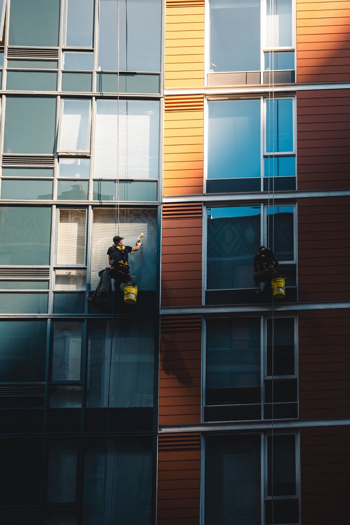 services-04 Two workers hanging to clean windows on a modern skyscraper facade.